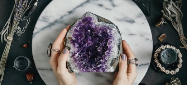 two hands holding half a purple geode on a marble base, with sage and candles around it
