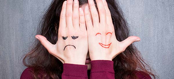 a woman covers her face with her hands that have both a smiley and a frownie face on them